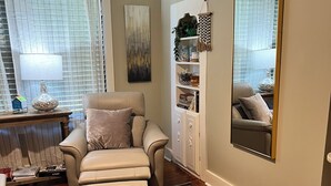 Una sala de estar con un sofá color beige, una mesa de centro de madera, un tapiz estampado, una estantería con objetos decorativos y un espejo grande.