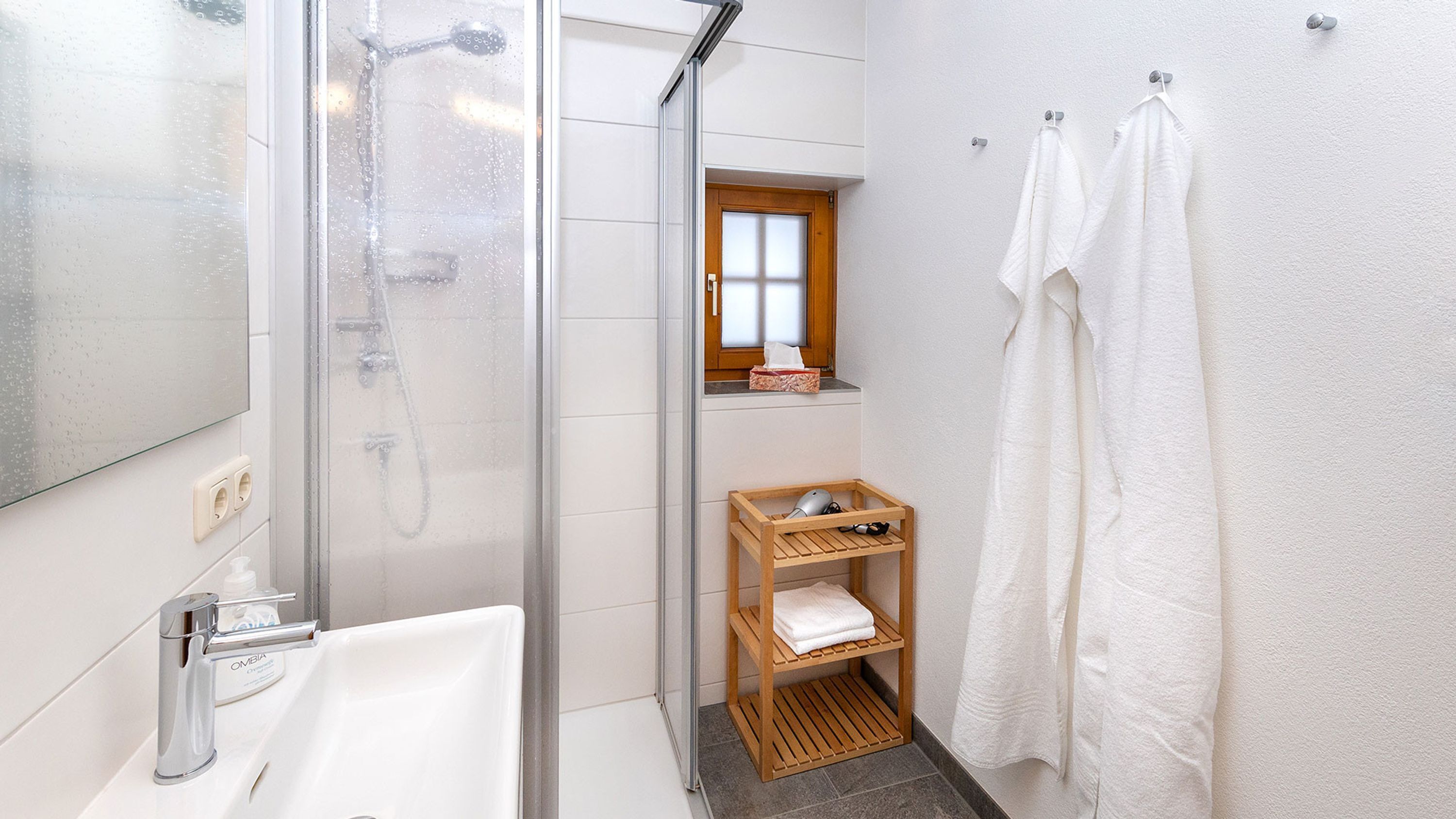 Un baño moderno con cabina de ducha de vidrio, un estante de madera con toallas y una pequeña ventana sobre el lavabo.