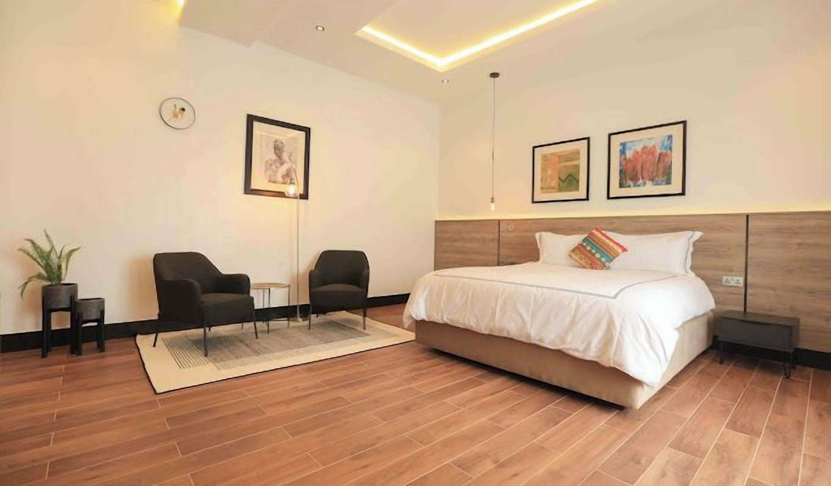 Une chambre d’hôtel moderne avec un grand lit, deux fauteuils, une petite table, une plante en pot et deux tableaux encadrés au mur.