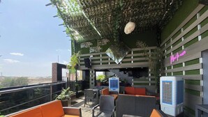 An outdoor seating area with wicker furniture and orange cushions, a small air conditioning unit, and a neon sign on the wall. - Piston Guest Inn (Kano)