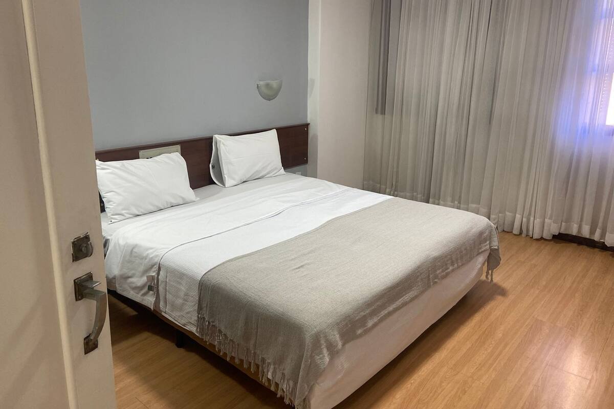 Une chambre d’h ôtel avec un lit, une table de chevet, une lampe fixée au mur et une fenêtre avec des rideaux légers.
