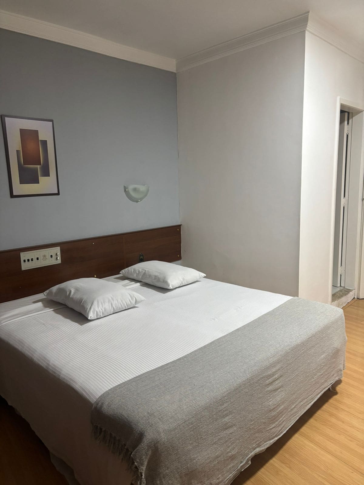 A neatly made bed with white linens and pillows, a wooden headboard, a bedside table, and a wall-mounted light fixture.