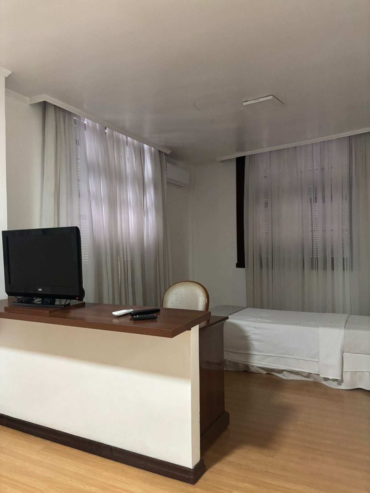 A hotel room with a wooden desk, a television, a chair, a bed, and sheer curtains.