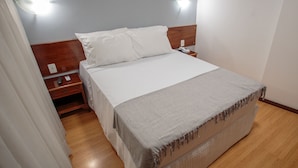 A neatly made bed with a white comforter and pillows, a wooden nightstand with a phone and charger, and a bedside lamp. - Fast 10 City Hotel (Porto Alegre)
