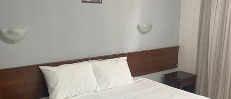 Una cama bien tendida con sábanas blancas, un cabecero de madera y una mesita de noche con una lámpara.