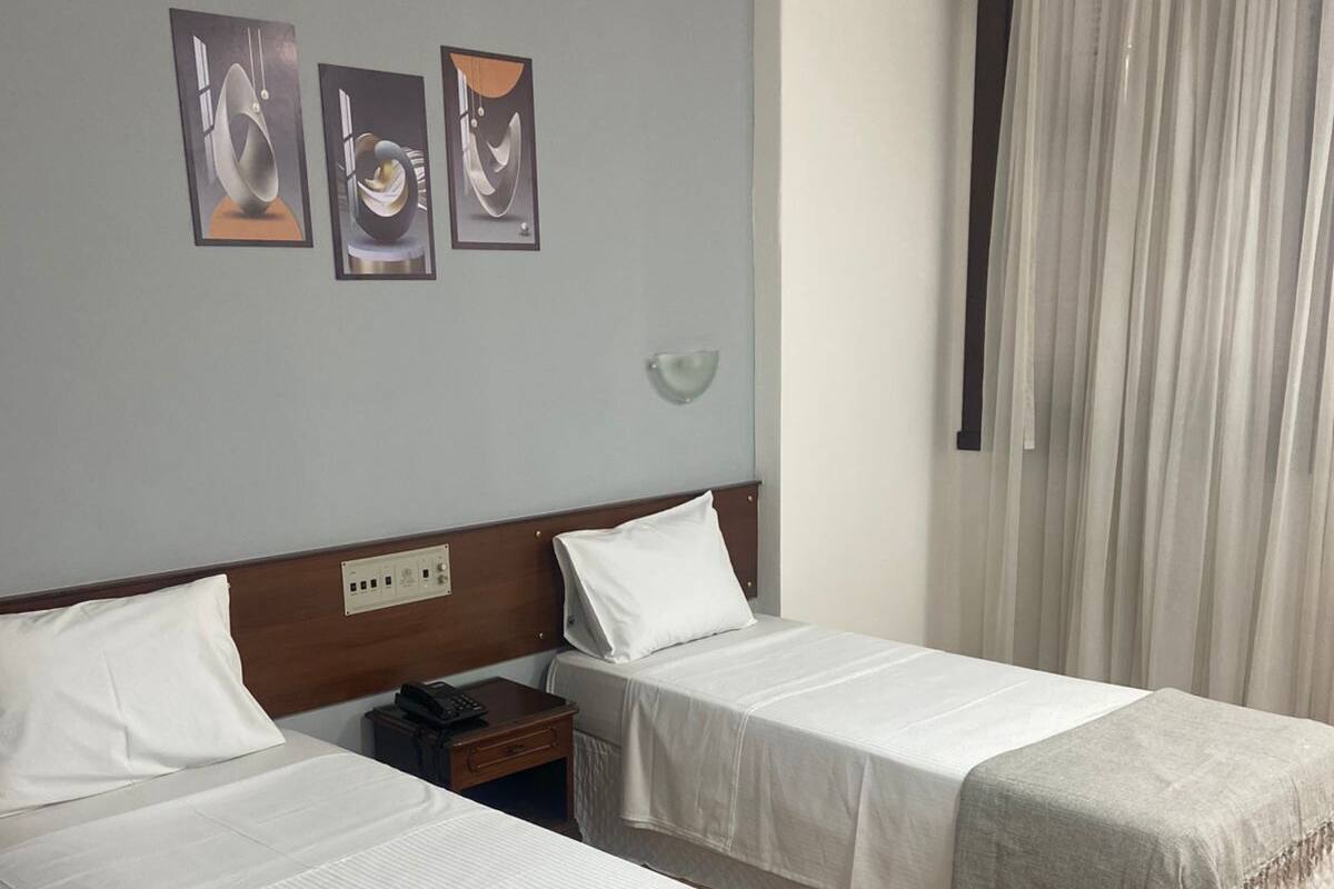 Une chambre d’hôtel avec deux lits simples, une table de chevet, une lampe fixée au mur et une fenêtre avec des rideaux.