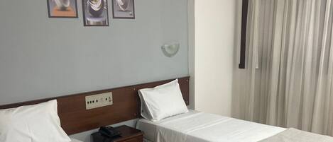 Habitación de hotel con dos camas individuales, una mesita de noche, una lámpara de pared y una ventana con cortinas.