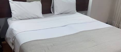 Una cama bien tendida con sábanas blancas y una manta de color beige, un cabecero de madera y una lámpara fijada a la pared.