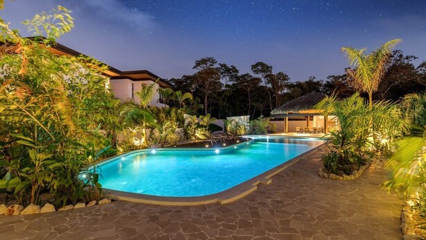 A swimming pool with a stone pathway, surrounded by lush greenery and a thatched-roof structure. - Casa Las Flores - Tropical Luxury Retreat Pinilla (Pinilla)