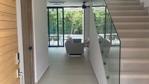 A modern interior with a glass staircase, a wooden wall, and a view of an outdoor area with a pool and furniture. - Casa Las Flores - Tropical Luxury Retreat Pinilla (Pinilla)