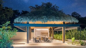 A modern outdoor dining area with a thatched roof, surrounded by greenery and under a starry night sky. - Casa Las Flores - Tropical Luxury Retreat Pinilla (Pinilla)