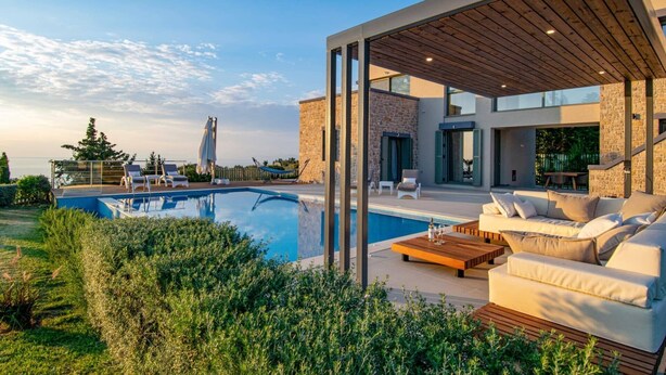 A modern outdoor pool area with a wooden deck, lounge chairs, and a stone building in the background.
