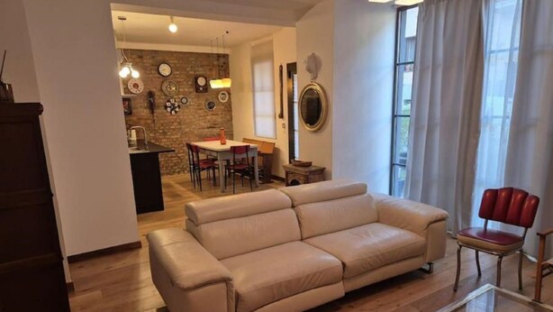 Un salon avec un canapé beige, une table basse en verre et un coin repas agrémenté d’un mur en briques et de luminaires suspendus.