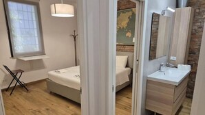 Une chambre à coucher comprenant un lit, une table de chevet, une lampe, une fenêtre et une vue sur une salle de bain avec un lavabo et un miroir.
