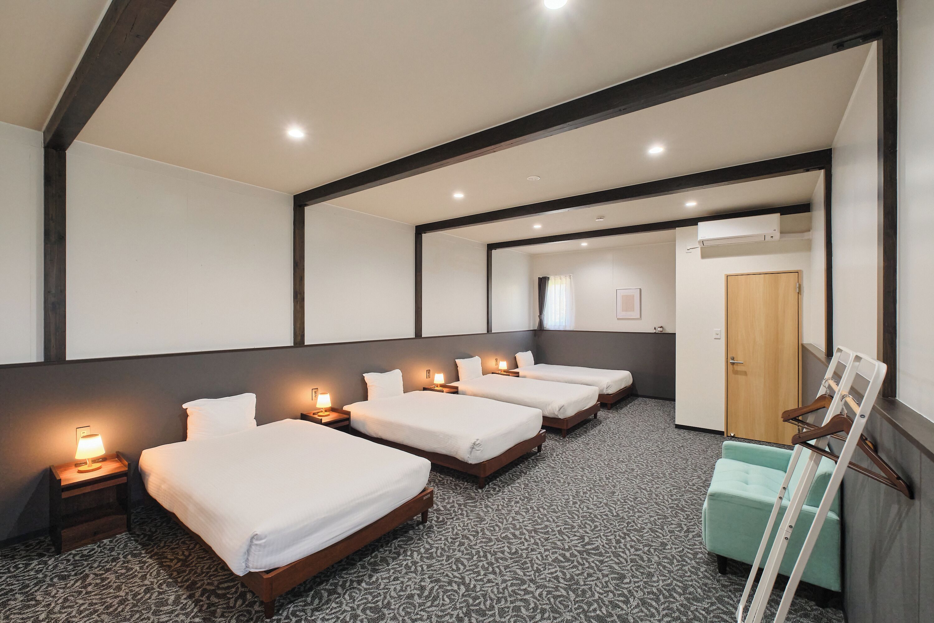 Habitación de hotel con cuatro camas individuales, una puerta de madera, un pequeño espacio de descanso y una alfombra estampada.