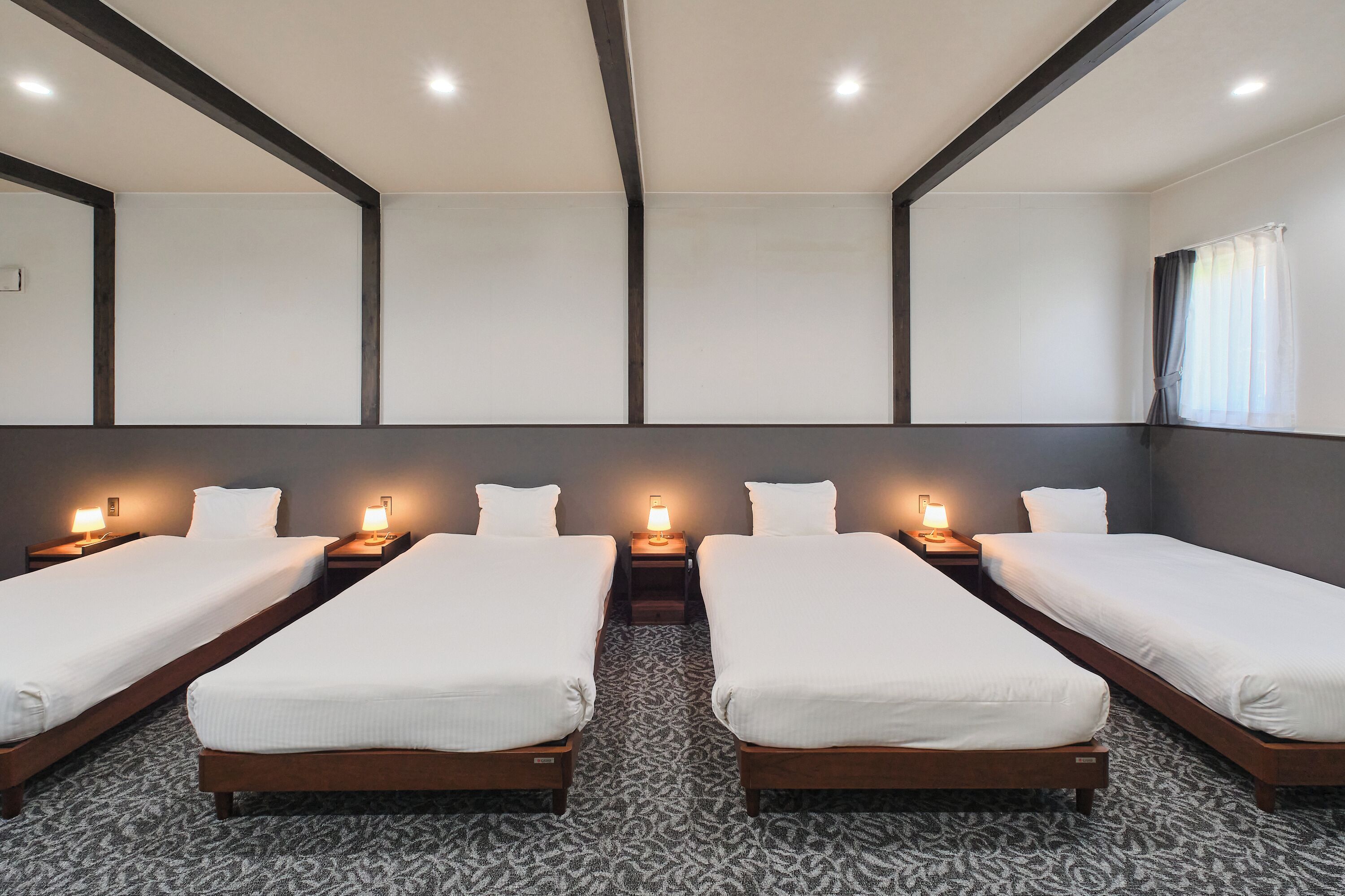 A hotel room with four single beds, each with a bedside table and lamp, and a large window with curtains.