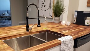 Una cocina con encimera de madera, pileta de acero inoxidable y muebles blancos. Hay una planta en la encimera y una cafetera.