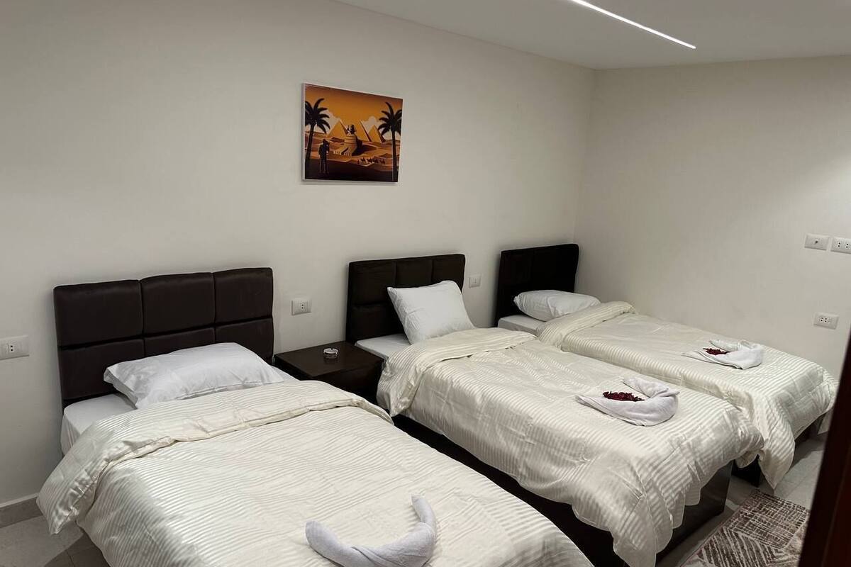 A hotel room with three single beds, each with a pillow and a towel. There is a painting on the wall above the beds.