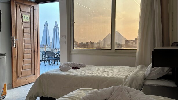Une chambre d’hôtel avec deux lits, une grande fenêtre donnant sur un paysage urbain et des montagnes, et un balcon avec des chaises et des parasols.