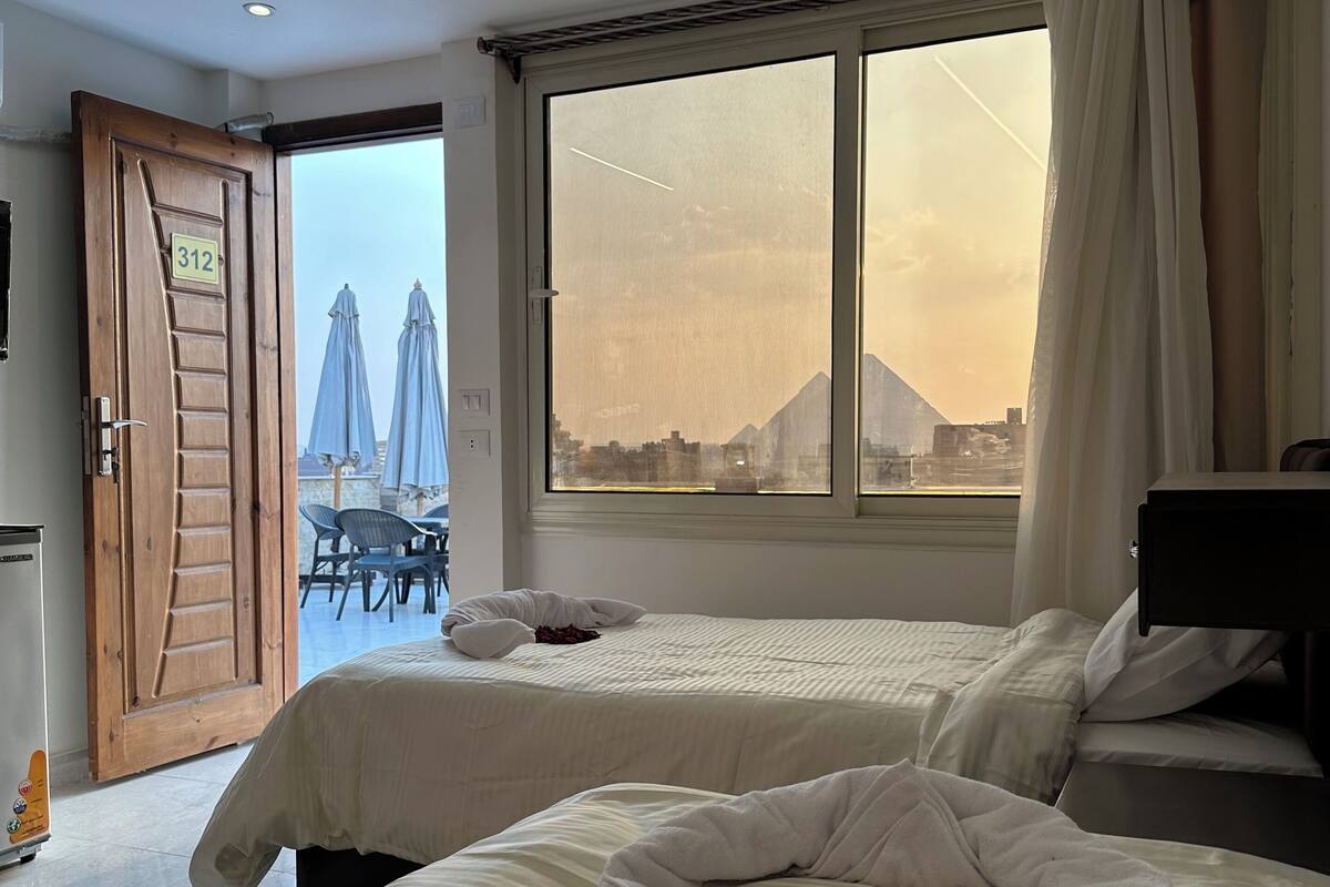 A hotel room with two beds, a large window with a view of a cityscape and mountains, and a balcony with chairs and umbrellas.