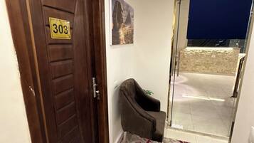 A hallway with a wooden door marked '303', a small armchair, a patterned rug, and a glass door leading outside.