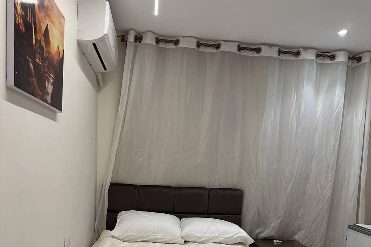 A neatly made bed with a dark headboard, a small bedside table with a lamp, and a wall-mounted air conditioner.