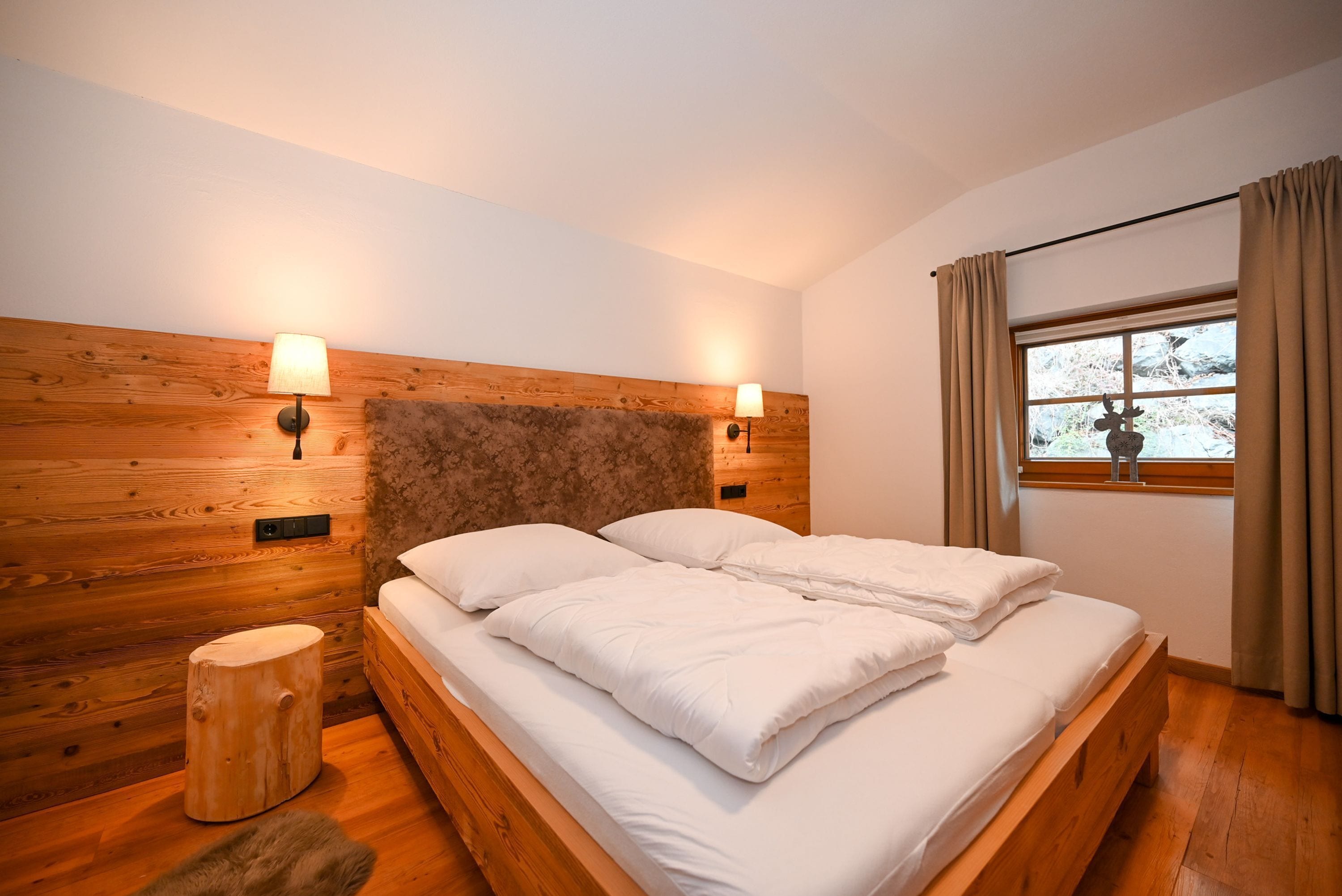 A bedroom with a wooden headboard, two bedside lamps, a window with curtains, and a small wooden stool.