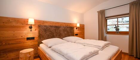 A bedroom with a wooden headboard, two bedside lamps, a window with curtains, and a small wooden stool.