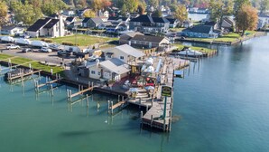 Un puerto deportivo con barcos amarrados, un pequeño edificio con un cartel, y casas al fondo.