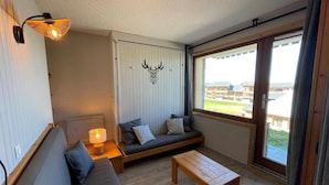 Ein gemütliches Zimmer mit einer Couch, einem Holztisch und einer Lampe. Es gibt ein großes Fenster mit Blick auf Gebäude.