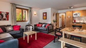 Un salon moderne avec un canapé gris, des coussins rouges et un tapis rouge. On y trouve une table à manger avec des chaises, ainsi qu’un coin cuisine doté de meubles en bois et d’un four à micro-ondes.
