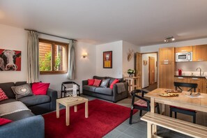 A modern living room with a grey sofa, red cushions, and a red rug. There is a dining table with chairs and a kitchen area with wooden cabinets and a microwave.