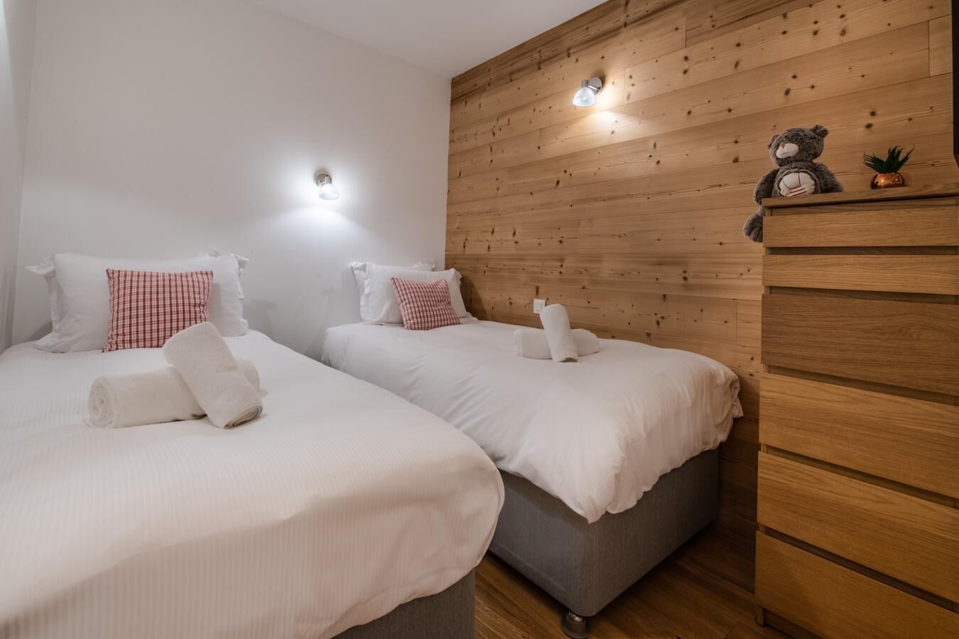 A hotel room with two beds, each with white linens and red and white checkered pillows. There are wall-mounted lights and a wooden dresser with a teddy bear and a plant on top.