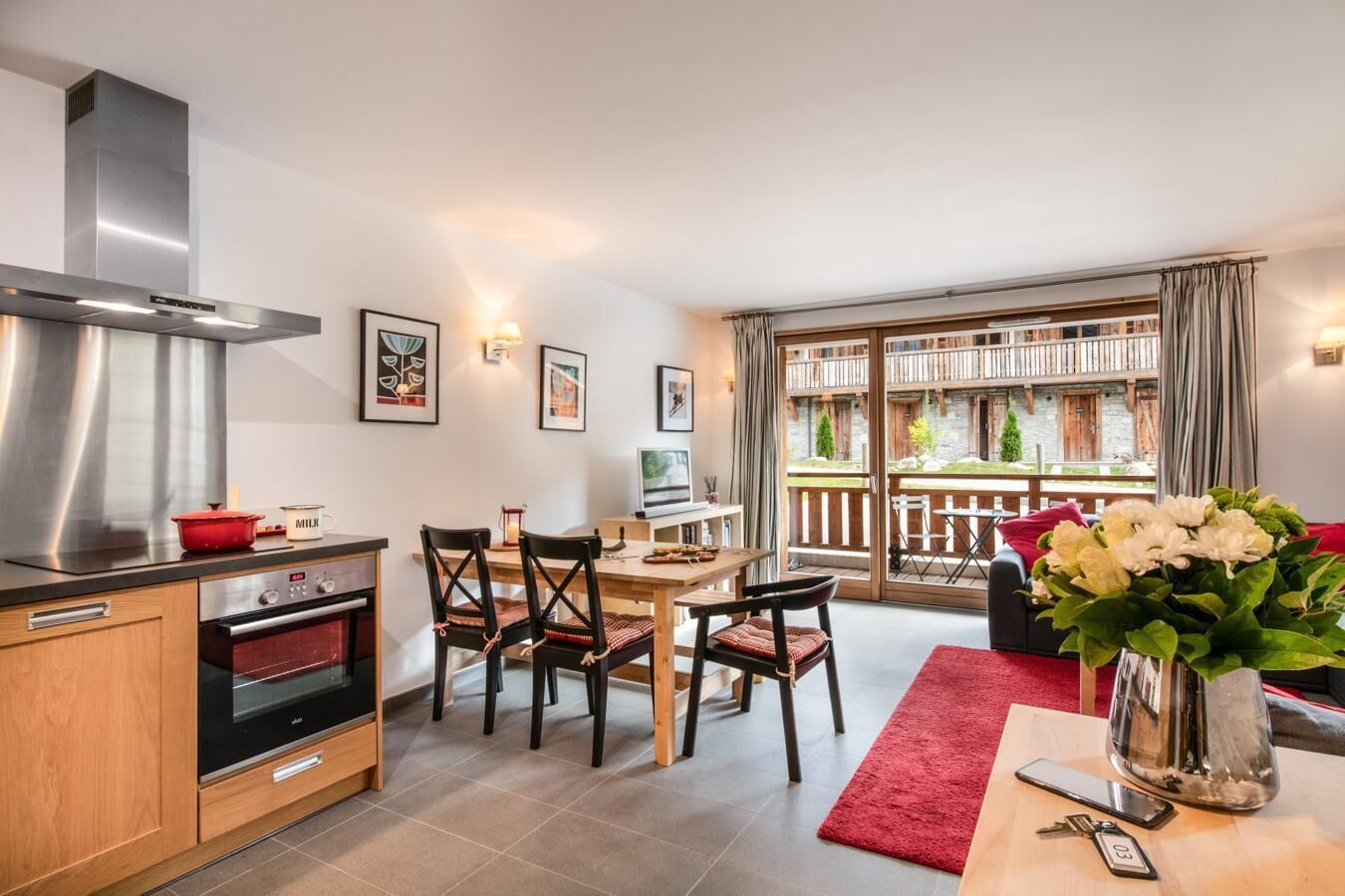 Une cuisine moderne équipée d’une cuisinière, d’un four et d’une hotte en acier inoxydable. On y trouve un coin repas avec une table et des chaises, ainsi qu’un tapis rouge. Un vase rempli de fleurs est posé sur une table au premier plan.