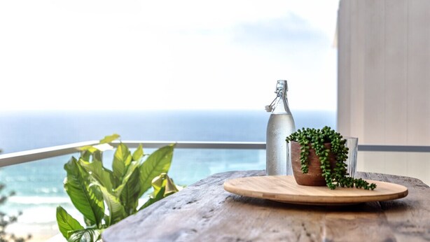 Une table en bois, une bouteille en métal, un verre et une plante en pot sont posés sur un balcon donnant sur l’océan.