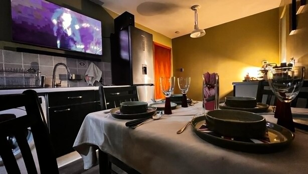 Un comedor con una mesa lista para dos, un televisor montado en la pared y una cocina al fondo.