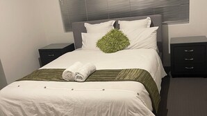 Una cama bien hecha con una colcha blanca y detalles verdes, un cabecero gris y una mesita de noche pequeña con cajones.