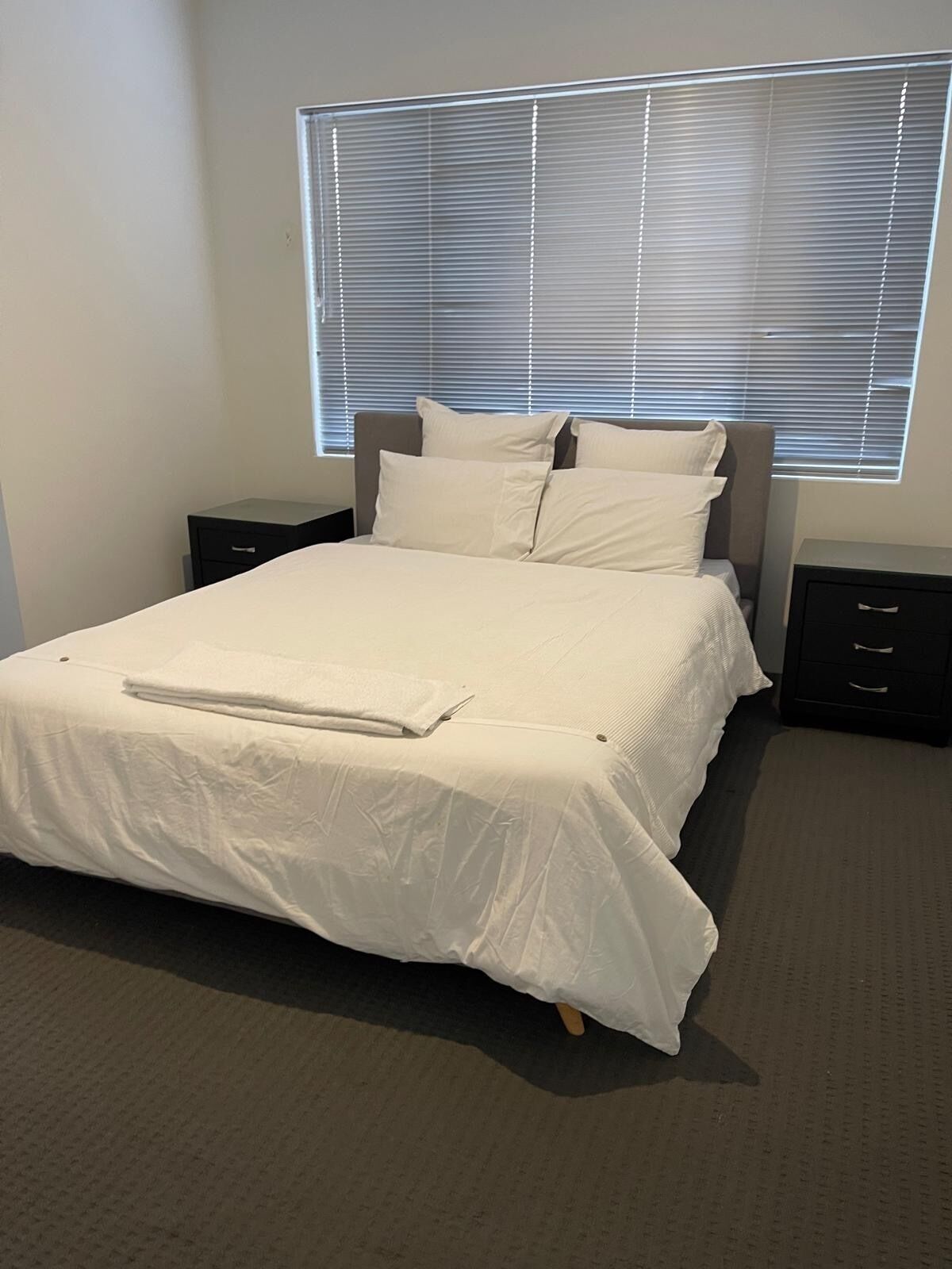 A neatly made bed with a white comforter and pillows, a bedside table with a lamp, and a window with blinds.