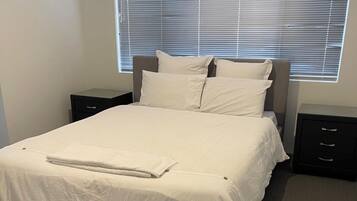 A neatly made bed with a white comforter and pillows, a bedside table with a lamp, and a window with blinds.