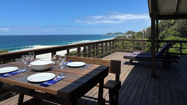 Una terraza de madera con una mesa dispuesta para dos, con vistas a una playa con olas y tierra en la distancia.