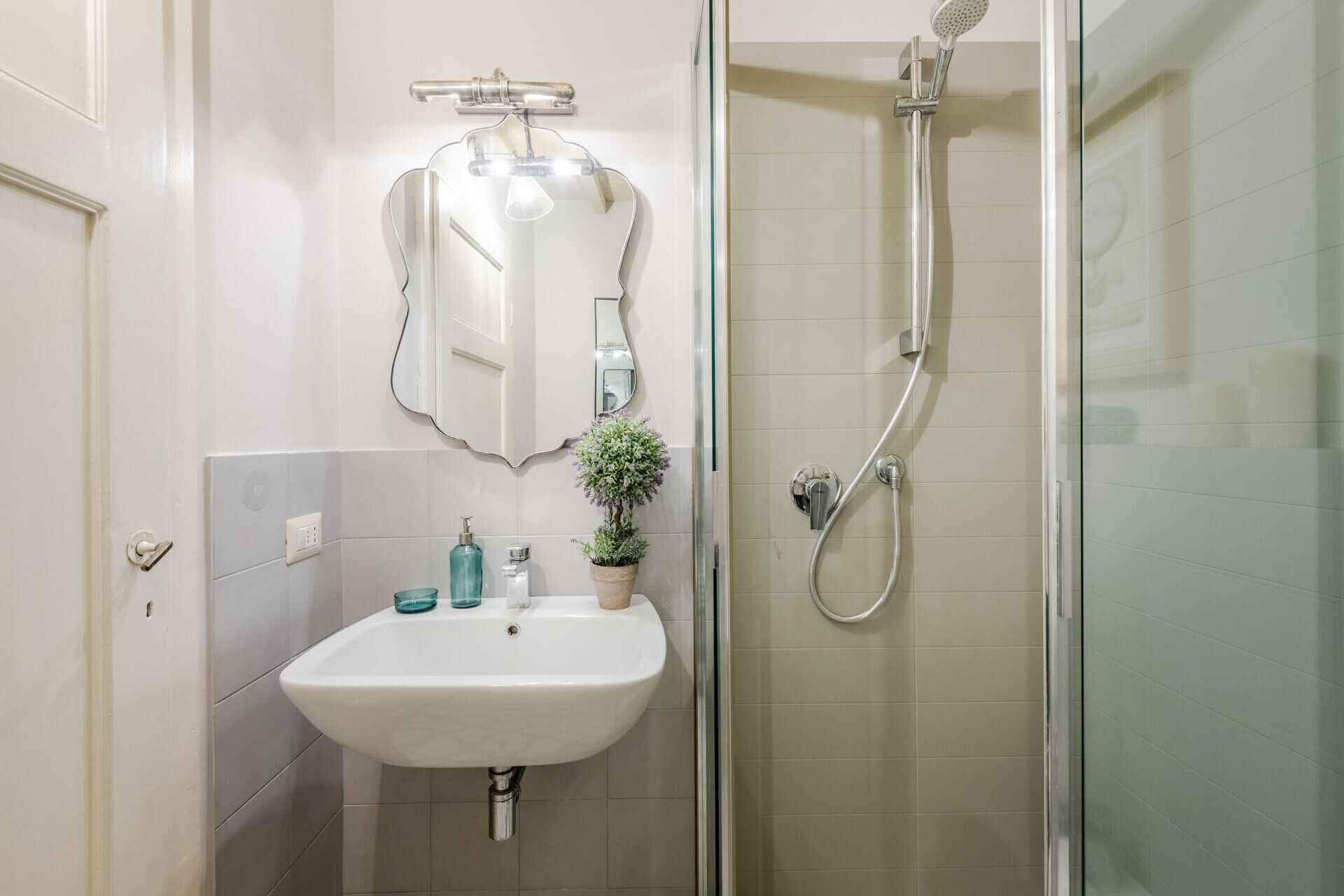 Ein Badezimmer mit weißem Waschbecken, einem Spiegel, einer Dusche und einer Topfpflanze.