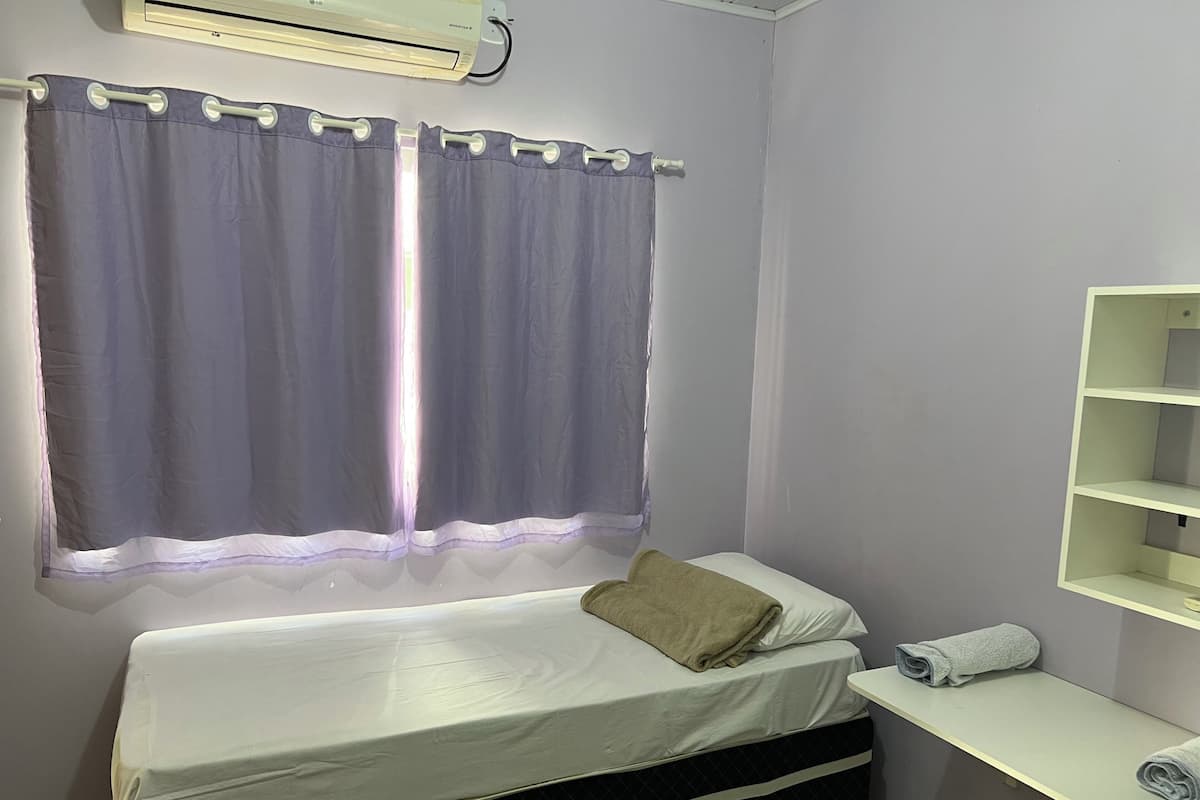 Une petite chambre à coucher, bien rangée, avec un lit simple, une fenêtre avec des rideaux, une étagère fixée au mur et un appareil de climatisation.