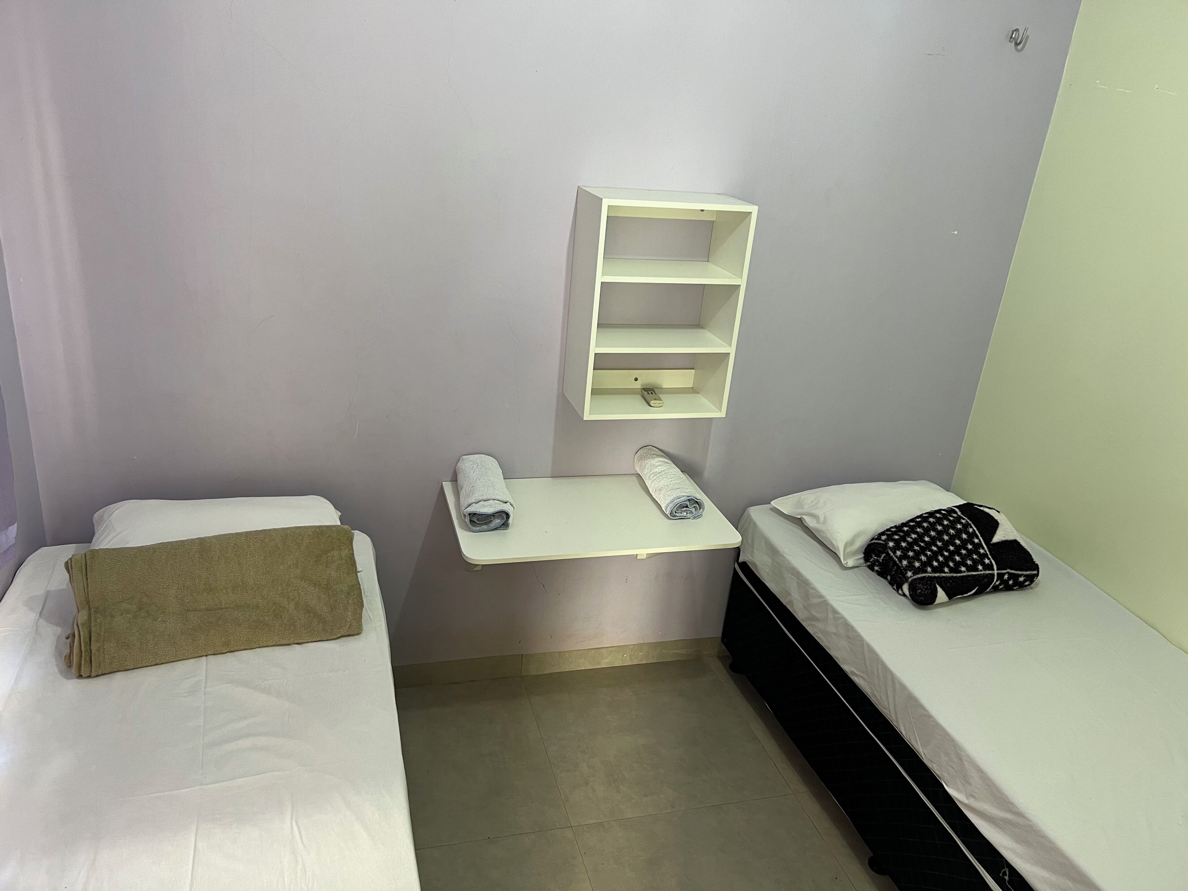 A small, clean room with two single beds, a small table with a towel, a shelf with a phone, and a wall-mounted light.