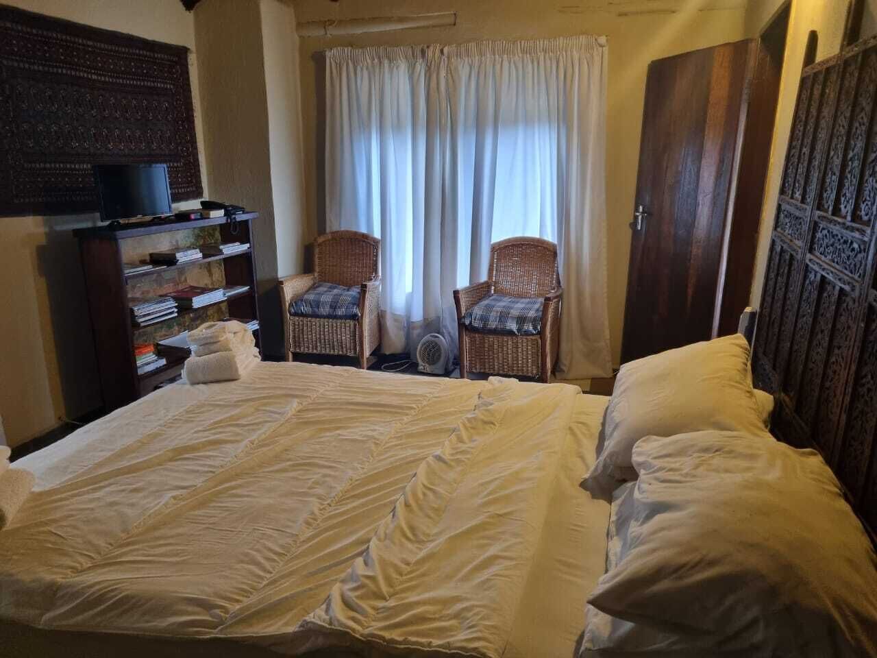 A bedroom with a large bed, two wicker chairs, a bookshelf, and a window with curtains.