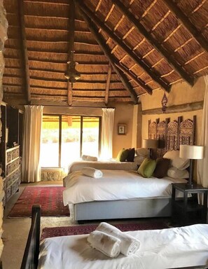 A room with a thatched roof, a bed with white linens, a bedside table with a lamp, and a view of the outdoors through a window.