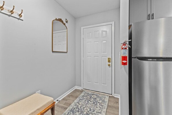 A hallway with a white door, a bench, a patterned rug, a stainless steel refrigerator, and a fire extinguisher.
