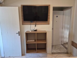 A room with a television mounted on the wall, a wooden console table, and a tiled floor. - Kitnet com ar na praia das Astúrias (Guarujá)
