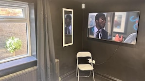 A room with a large window, a radiator, a white folding chair, and a wall-mounted TV displaying a scene. - Digz Hotel (Middlesbrough)
