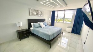 A modern bedroom with a bed, bedside tables, a nightstand, a ceiling light fixture, and a window with curtains. - Beachfront  2 Bdrm ~ Walk to Eat ~ W/D ~ King Bed (Carolina)