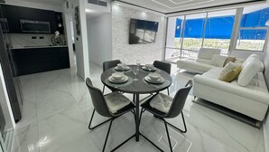 A modern living room with a dining area, a white sofa, and a flat-screen TV mounted on the wall. - Beachfront  2 Bdrm ~ Walk to Eat ~ W/D ~ King Bed (Carolina)
